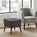 A square ottoman with a black and white geometric trellis patterned fabric top and dark wood legs, styled next to a light gray armchair in a bright room with a window.