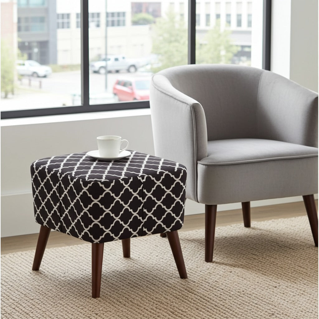 A square ottoman with a black and white geometric trellis patterned fabric top and dark wood legs, styled next to a light gray armchair in a bright room with a window.