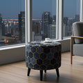 A round upholstered ottoman or small table with a dark blue and gold geometric hexagon pattern, supported by four black legs, set in a modern room with a city skyline view through large windows.
