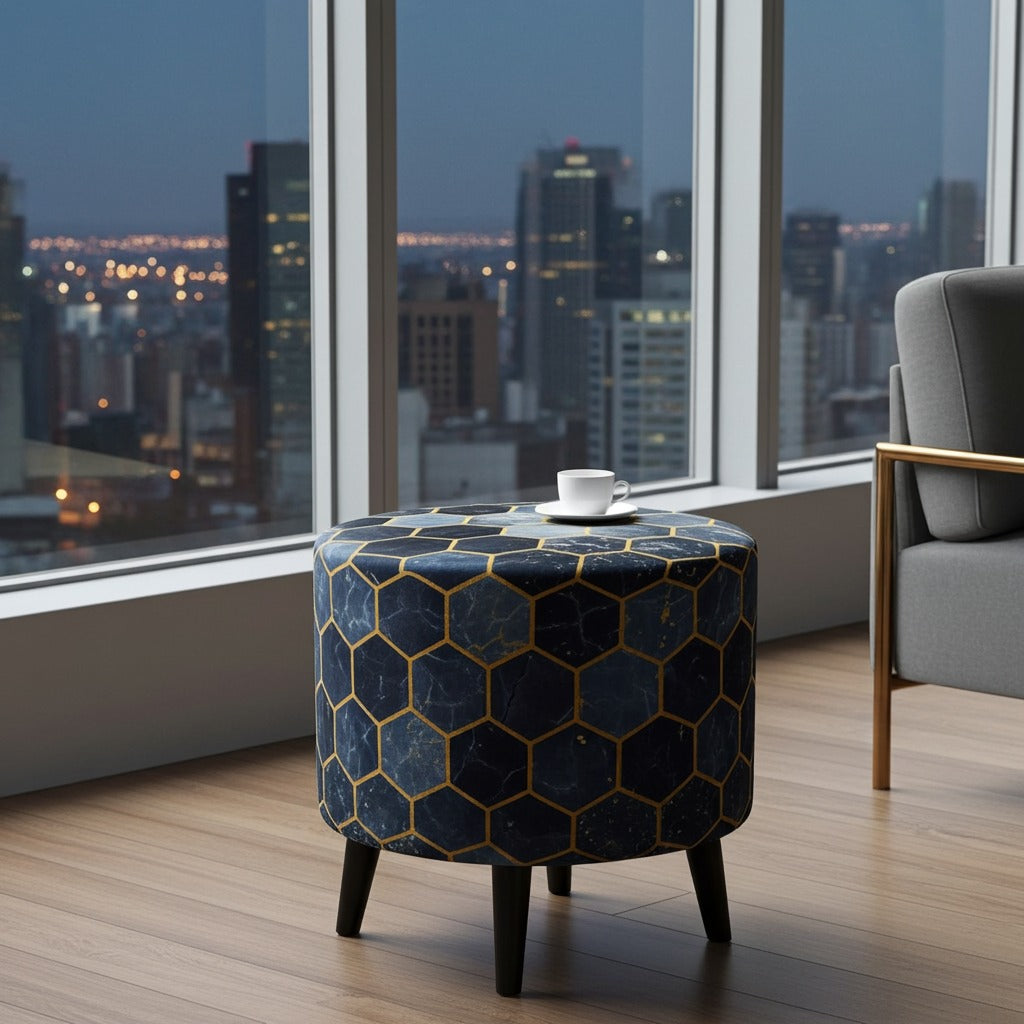 A round upholstered ottoman or small table with a dark blue and gold geometric hexagon pattern, supported by four black legs, set in a modern room with a city skyline view through large windows.
