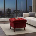 A tufted red velvet storage ottoman with wooden legs sits on a light beige rug in a modern living room setting with a city skyline at dusk visible through large windows.