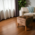 A brown tufted velvet ottoman with wooden legs in a modern living room setting with a beige sofa, white curtains, and a potted plant.