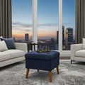 A navy blue tufted ottoman with light wood legs in a modern living room setting, positioned in front of large windows showing a city skyline at dusk.