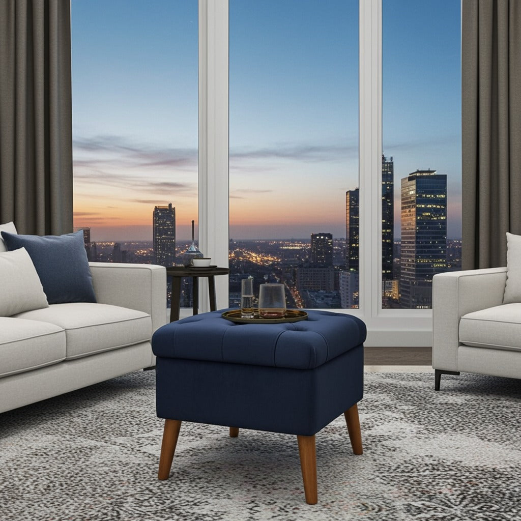 A navy blue tufted ottoman with light wood legs in a modern living room setting, positioned in front of large windows showing a city skyline at dusk.