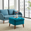 A bright blue upholstered loveseat with matching pillows sits in a modern living room next to a matching tufted ottoman with wooden legs. Large windows and a light rug are visible in the background.