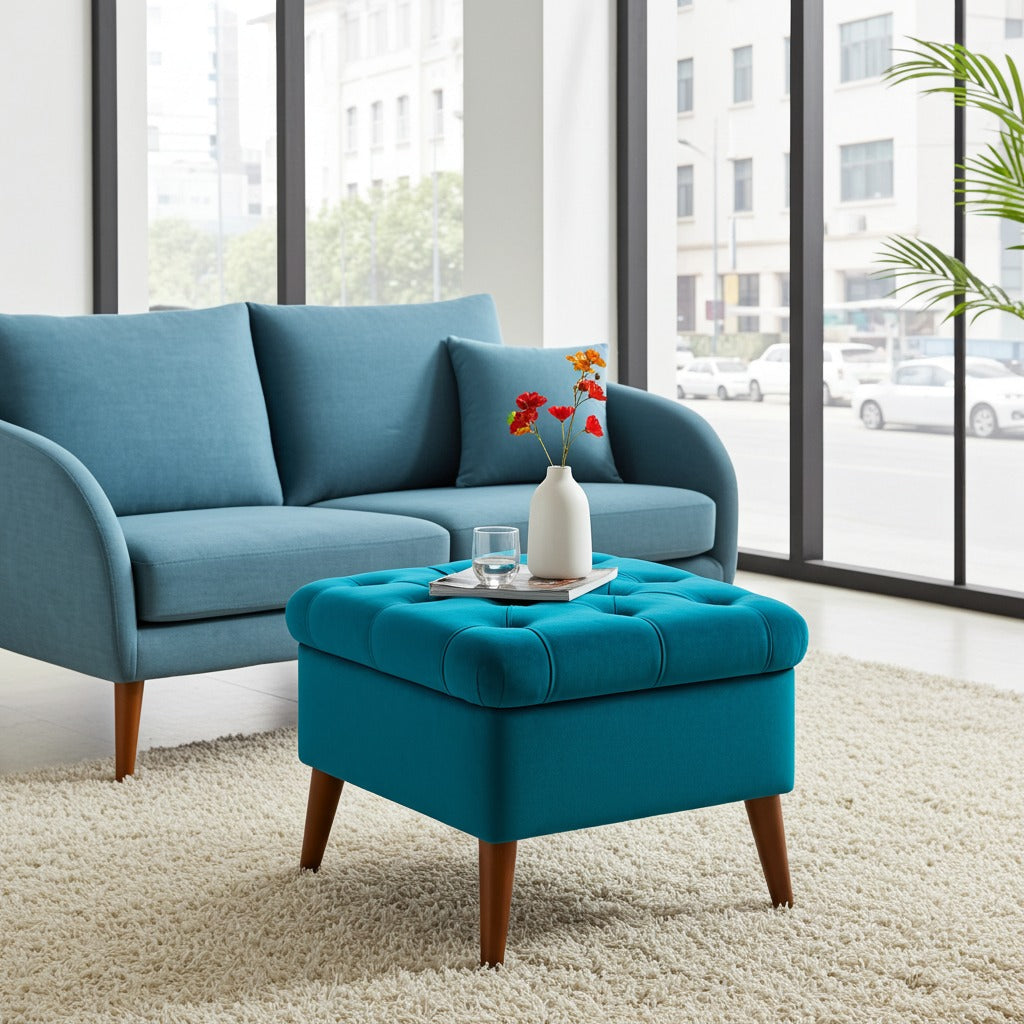 A bright blue upholstered loveseat with matching pillows sits in a modern living room next to a matching tufted ottoman with wooden legs. Large windows and a light rug are visible in the background.