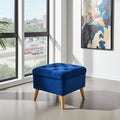 A blue tufted storage ottoman with light wood legs, positioned in a modern room next to a large window with an outdoor view.