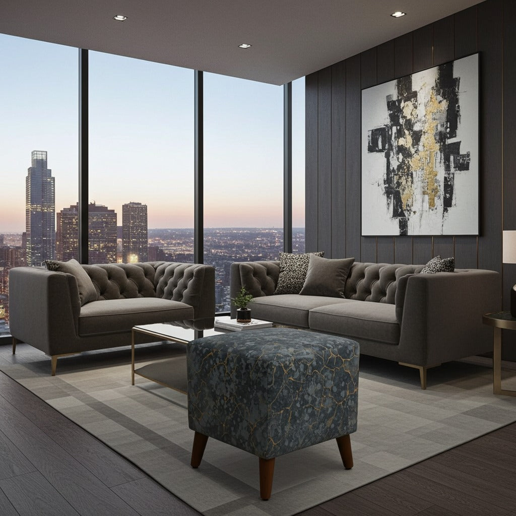 A modern living room setup featuring a tufted gray sofa and matching armchair with a blue patterned ottoman, set in front of a large window overlooking a city skyline at dusk.
