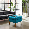 A bright turquoise blue tufted ottoman with tapered wooden legs, styled in a modern living room setting next to a black leather sofa and large potted plants.
