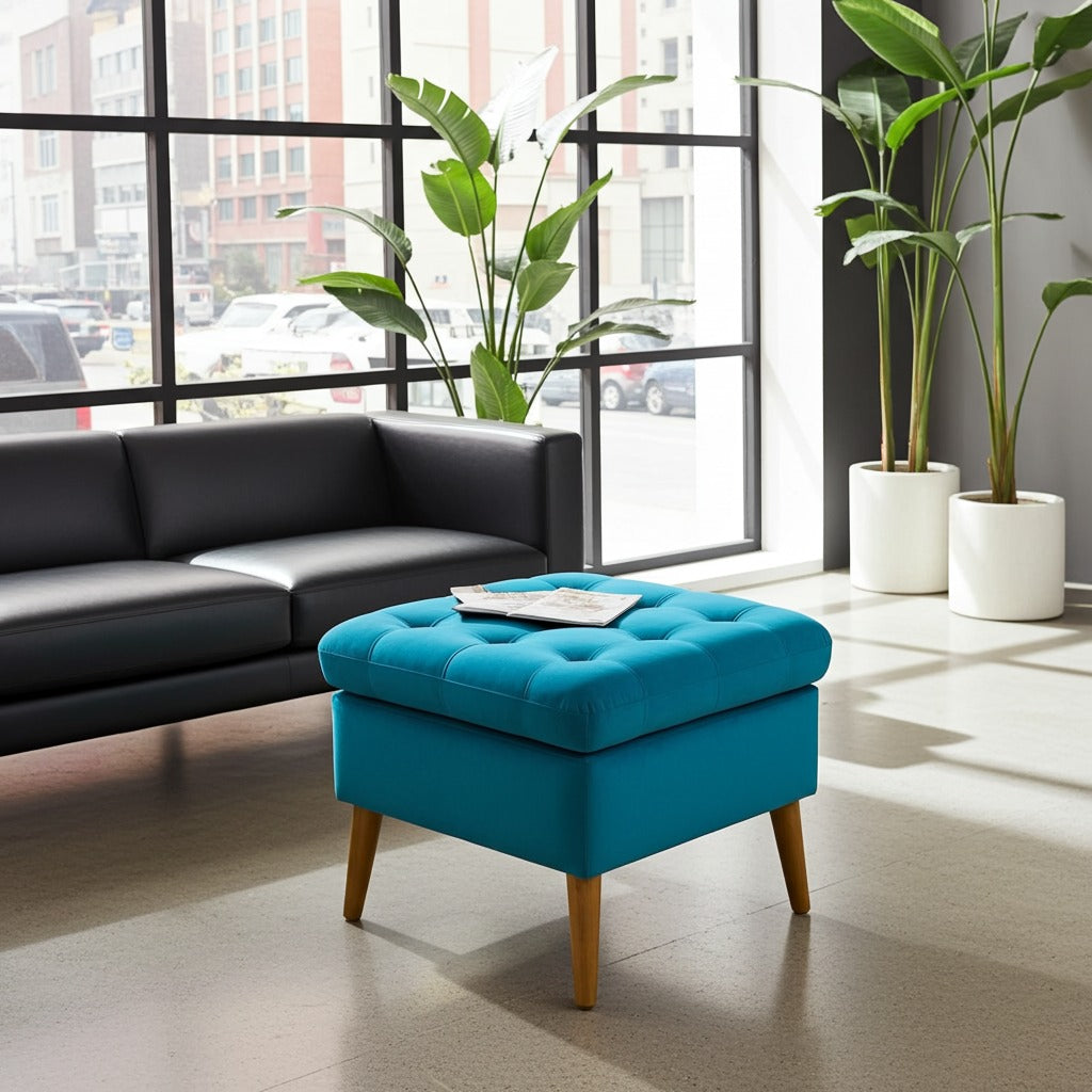 A bright turquoise blue tufted ottoman with tapered wooden legs, styled in a modern living room setting next to a black leather sofa and large potted plants.