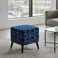 A square velvet ottoman in deep blue with a geometric gold line pattern and dark tapered legs, styled in a modern living room next to a glass-top side table and a gray armchair.