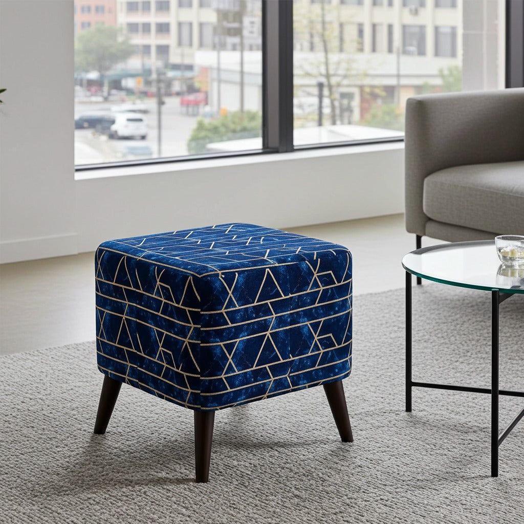 A square velvet ottoman in deep blue with a geometric gold line pattern and dark tapered legs, styled in a modern living room next to a glass-top side table and a gray armchair.