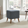 A square upholstered ottoman stool with a gray and blue geometric patterned fabric and tapered wooden legs, positioned on a white rug in a bright living room near a window.