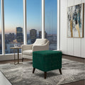 A modern living room scene featuring a deep emerald green velvet ottoman with dark wooden legs in the foreground, next to a white armchair and a small side table, all set on a patterned gray rug in front of large floor-to-ceiling windows showing a city skyline at dusk.