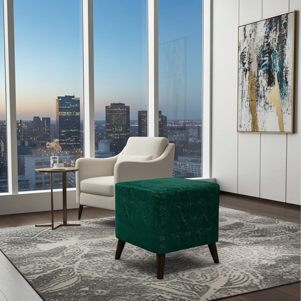 A modern living room scene featuring a deep emerald green velvet ottoman with dark wooden legs in the foreground, next to a white armchair and a small side table, all set on a patterned gray rug in front of large floor-to-ceiling windows showing a city skyline at dusk.