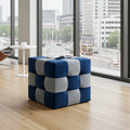 A cube-shaped tufted ottoman in a blue and light gray checkered pattern, placed on a light wood floor in a modern room with large windows overlooking a city street.