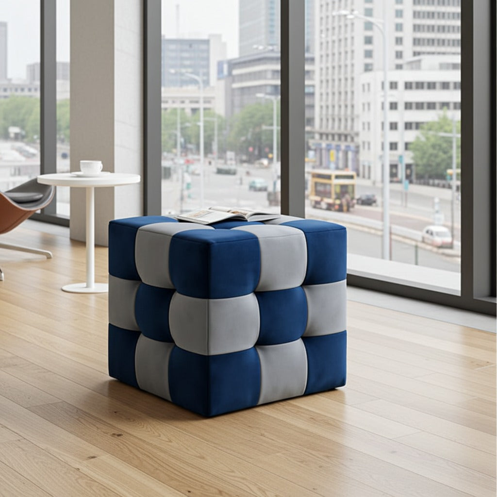 A cube-shaped tufted ottoman in a blue and light gray checkered pattern, placed on a light wood floor in a modern room with large windows overlooking a city street.