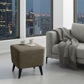 A square upholstered ottoman in taupe-brown fabric with dark tapered legs stands beside a light gray sofa in a modern room with large windows overlooking a city skyline at dusk. Two white mugs sit on the ottoman.