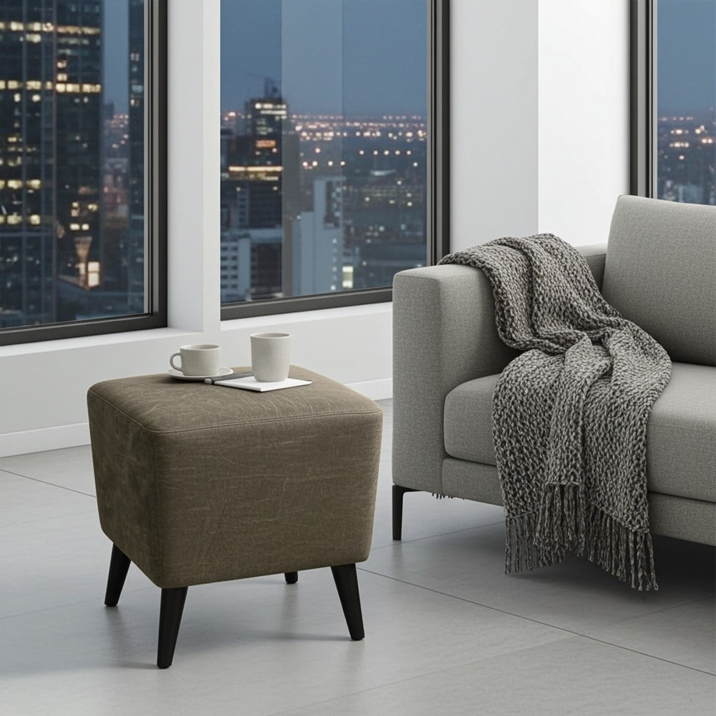 A square upholstered ottoman in taupe-brown fabric with dark tapered legs stands beside a light gray sofa in a modern room with large windows overlooking a city skyline at dusk. Two white mugs sit on the ottoman.