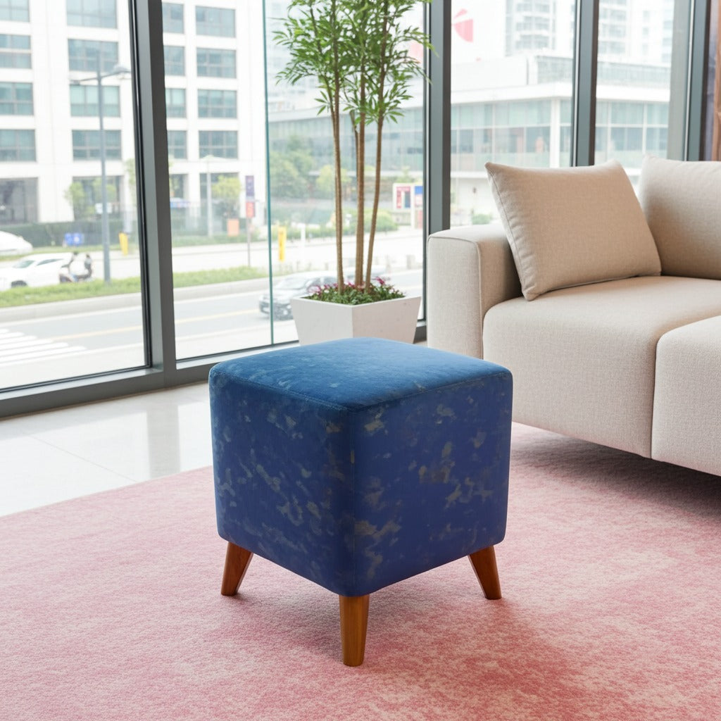 A square blue velvet ottoman with a subtle gold pattern and wooden legs, styled in a modern living room next to a beige sofa on a pink rug.