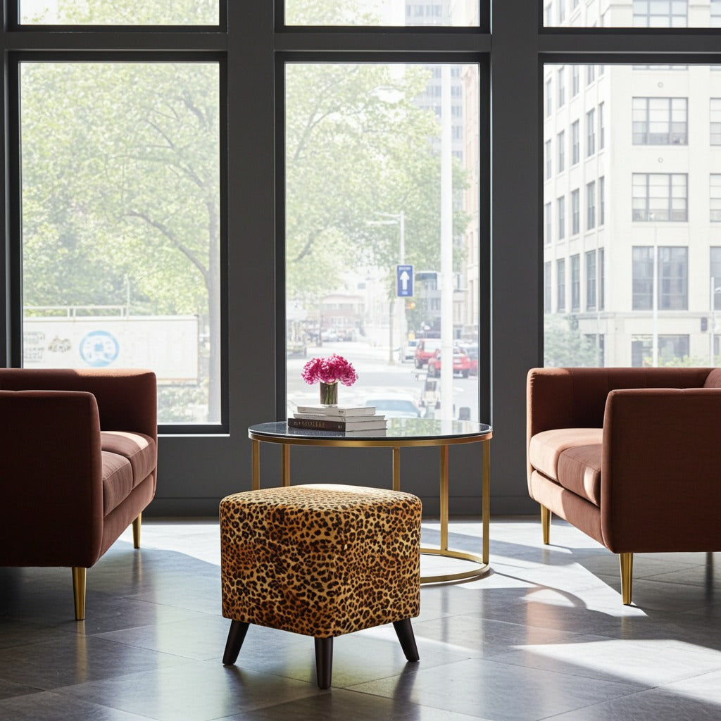 A modern lounge area with two rust-brown armchairs and a leopard-print ottoman around a round glass coffee table with gold legs, set in front of large windows overlooking a city street.