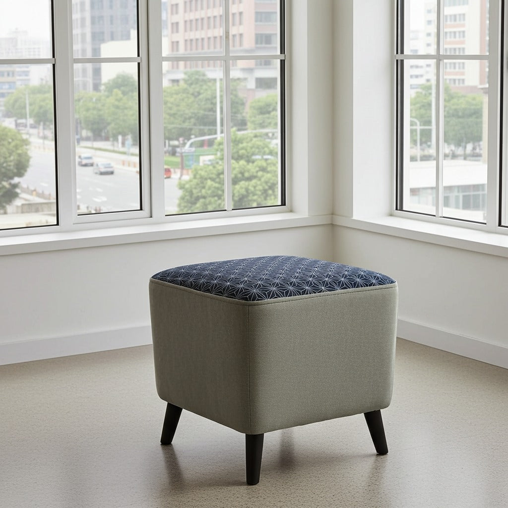 A square upholstered ottoman stool with a light beige fabric body, a dark blue patterned tufted top, and dark tapered legs, positioned in a bright room with large windows showing an outdoor city view.