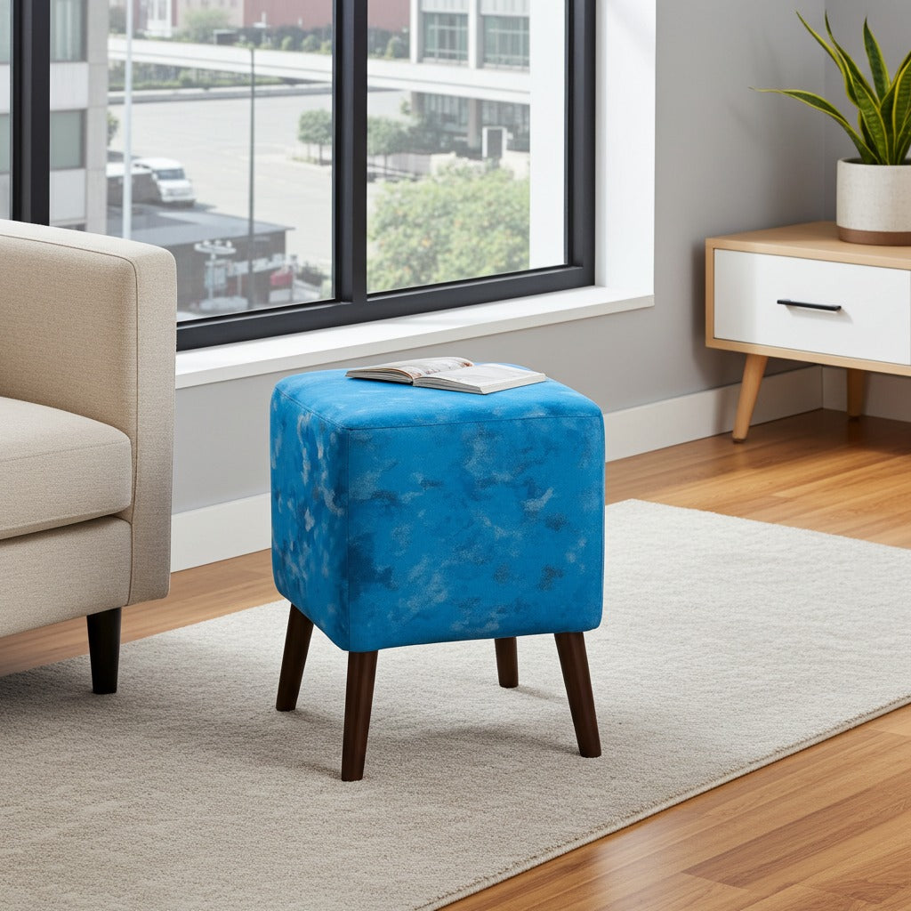 A square ottoman stool with a bright blue marbled velvet upholstery and dark wood legs, styled in a modern living room setting next to a beige sofa and a wooden side table.