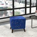 A square blue velvet ottoman with a distressed texture and dark wooden legs, positioned on a light gray rug in front of a large window with a city view.
