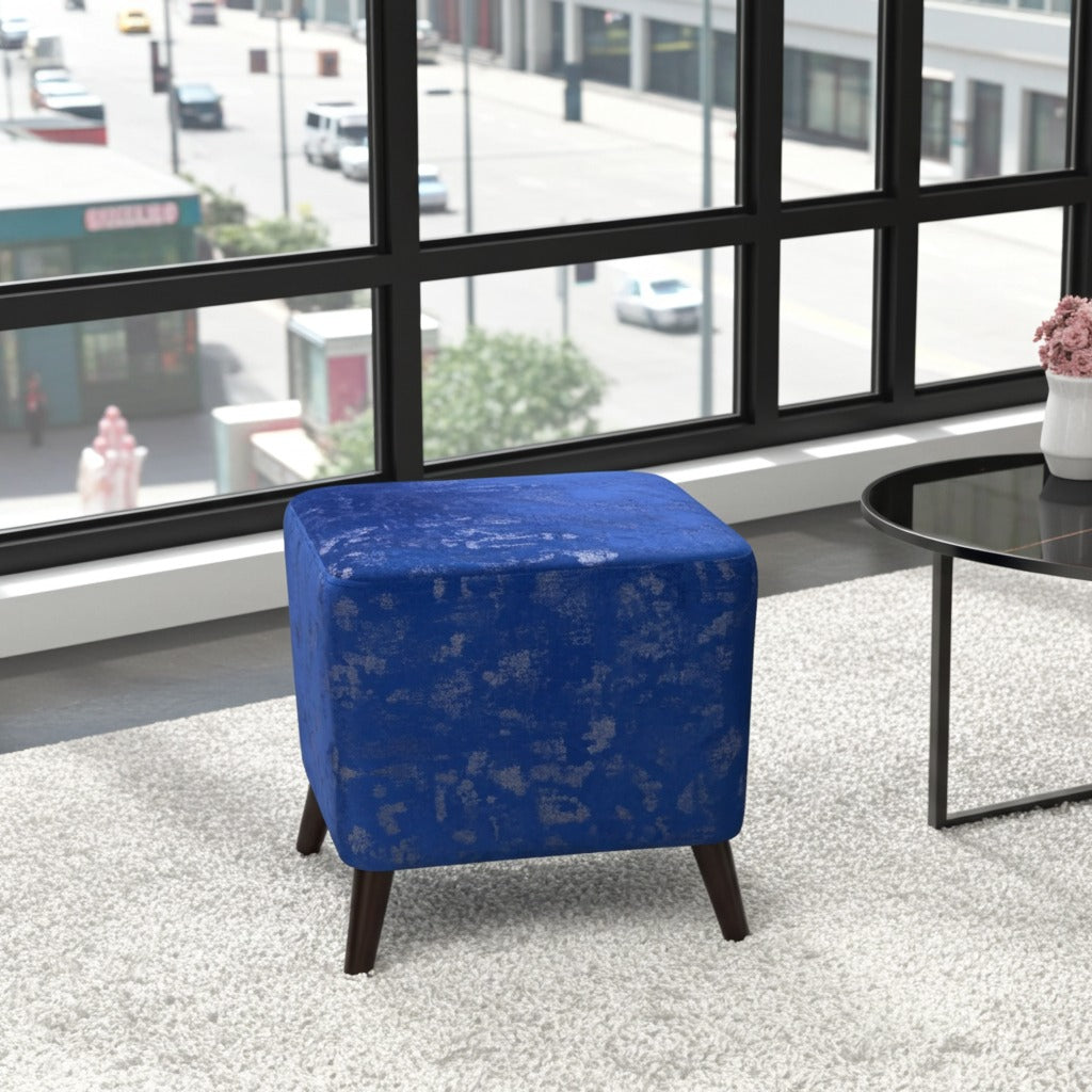 A square blue velvet ottoman with a distressed texture and dark wooden legs, positioned on a light gray rug in front of a large window with a city view.
