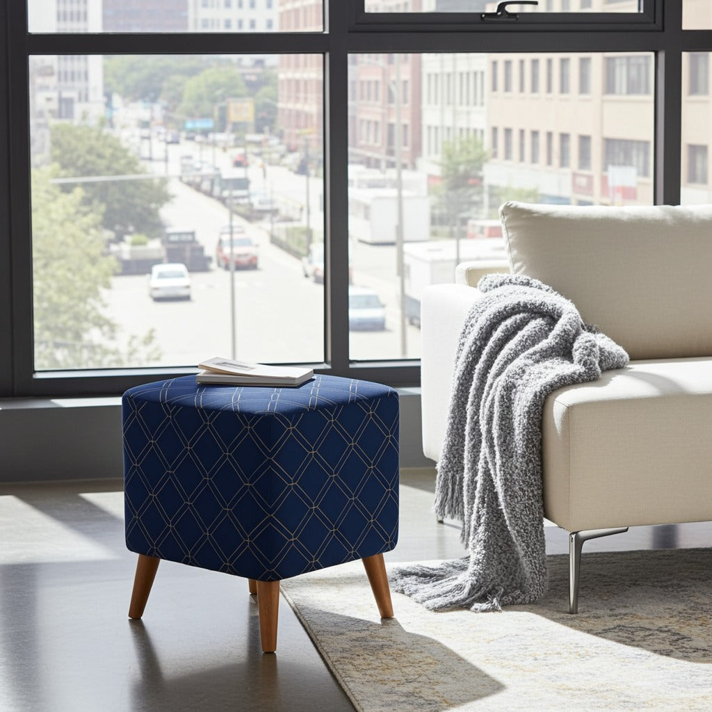 A blue upholstered ottoman with a geometric diamond pattern and wooden legs, styled in a modern living room next to a white armchair with a gray throw blanket, in front of a large window showing a city street view.
