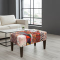 A square ottoman with a colorful patchwork print and dark brown wooden legs, styled in a modern living room setting next to a white sofa and a glass coffee table.
