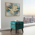 A teal velvet upholstered ottoman with a gold geometric pattern and dark wooden legs, styled in a modern room next to a large window showing a city skyline. An abstract teal and gold painting hangs on the wall above.