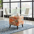 A square upholstered ottoman with a vibrant orange, red, and beige floral pattern sits on a blue patterned rug in a modern room with large windows, white chairs, and a city view.
