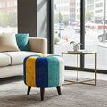 A colorful upholstered ottoman stool with vertical panels in blue, yellow, teal, and off-white, supported by dark wooden legs, shown in a modern living room next to a side table.