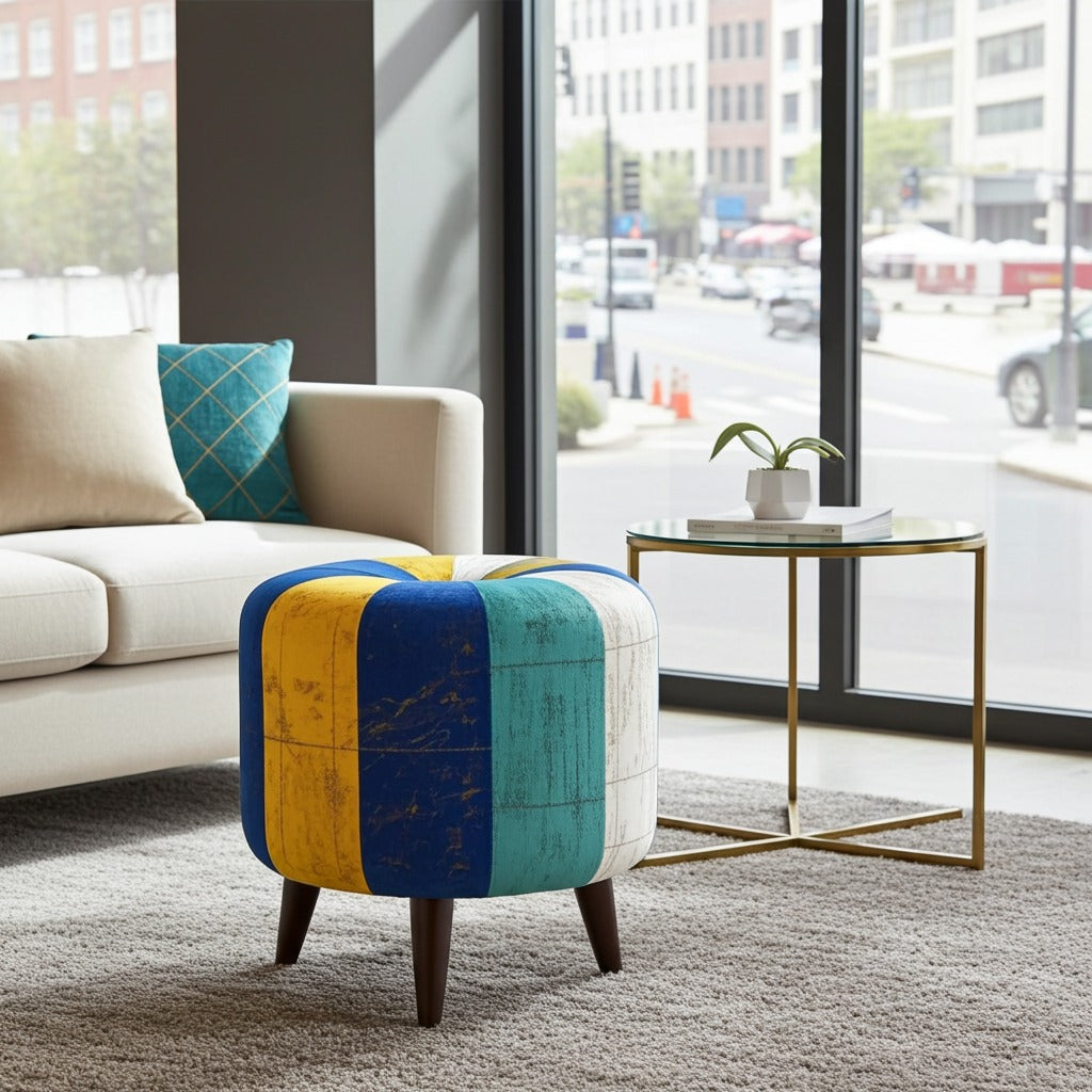 A colorful upholstered ottoman stool with vertical panels in blue, yellow, teal, and off-white, supported by dark wooden legs, shown in a modern living room next to a side table.