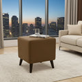 A brown upholstered ottoman with dark tapered legs sits on a white rug in a modern living room, with a city skyline visible through large windows behind it.