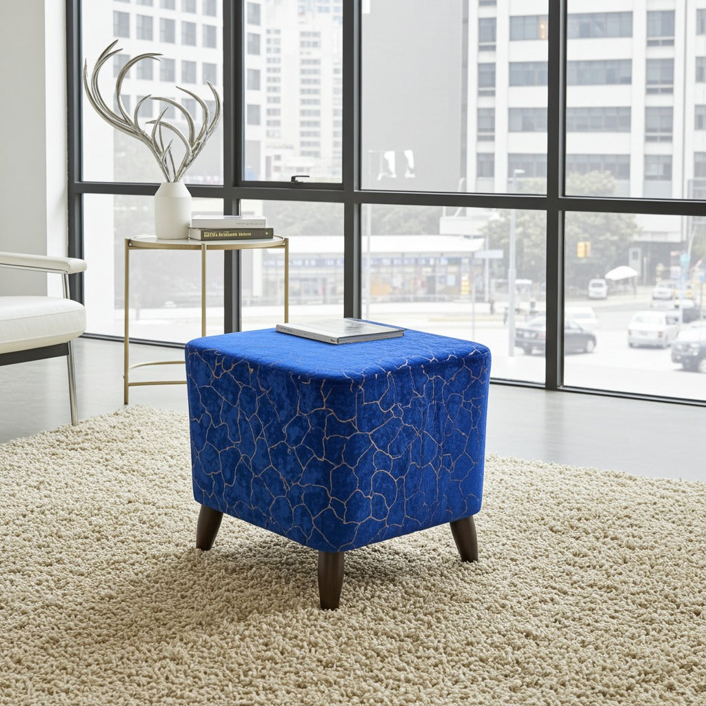 A square blue velvet ottoman with a gold geometric crackle pattern and dark wooden legs, styled on a beige shag rug in a modern room with large windows.