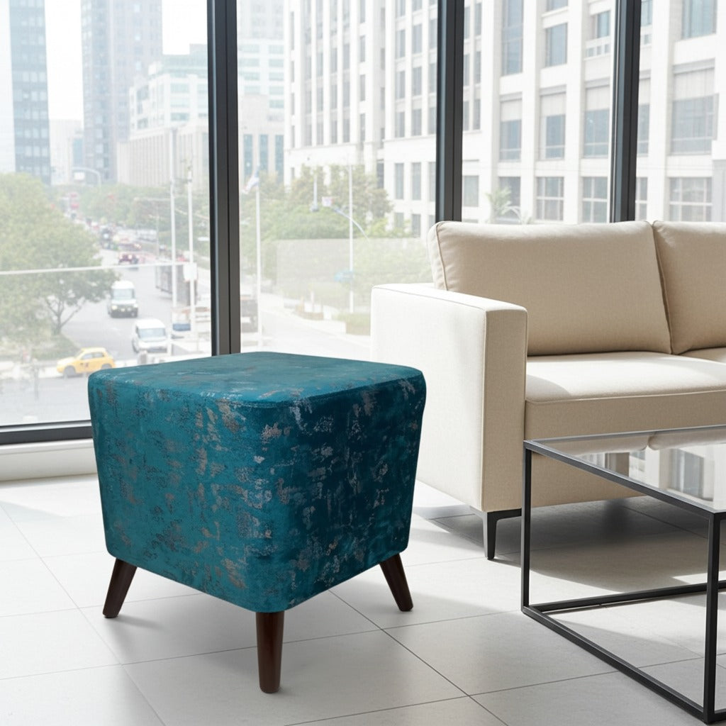 A square teal velvet ottoman with a subtle floral pattern and dark wooden legs, styled in a modern living room next to a beige sofa and a glass-top coffee table.