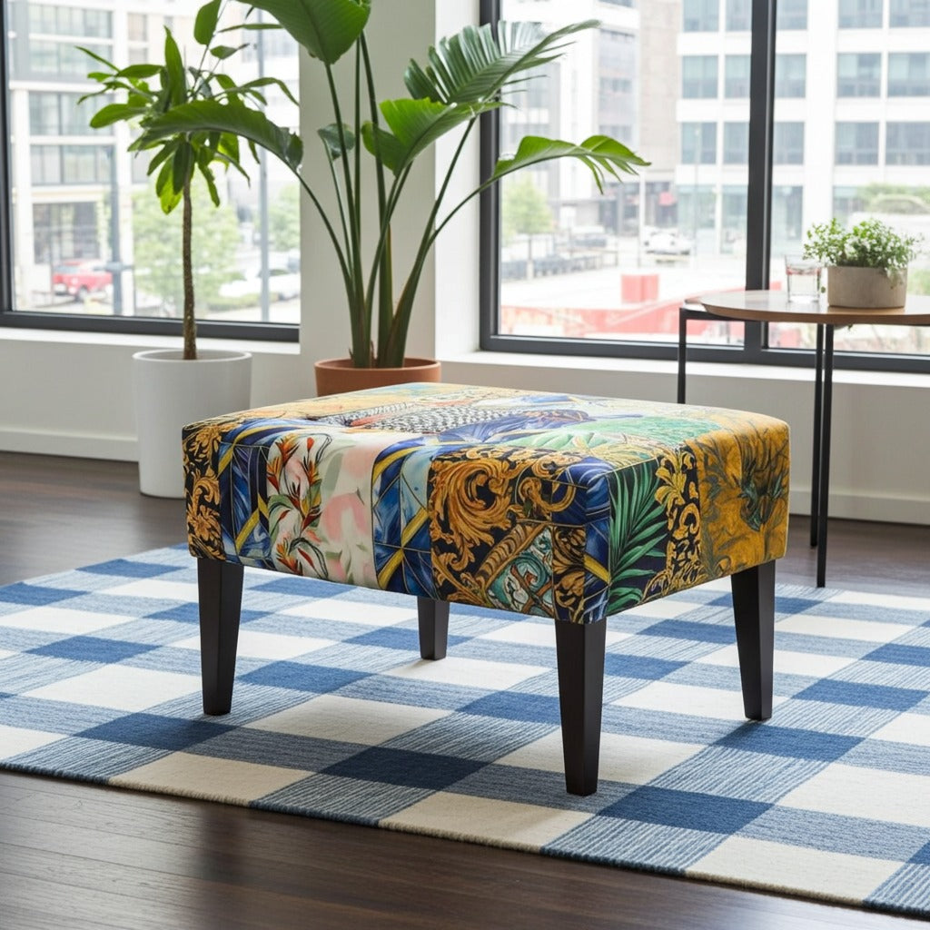 A square upholstered ottoman stool with a vibrant multicolor baroque and botanical pattern, supported by four dark wooden legs, set on a blue and white checkered rug in a modern living room.