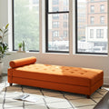 An orange tufted chaise lounge daybed with a matching cylindrical lumbar pillow, positioned on a patterned rug in a bright room with large windows overlooking a city street.