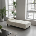 A cream-colored tufted leather chaise lounge with dark wooden legs, positioned in a modern, minimalist room with large windows, potted plants, and a gray concrete floor.