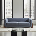 A two-seat sofa upholstered in textured blue fabric with dark wood legs, positioned in a modern living room setting with a glass-top coffee table and large windows showing a city street view.