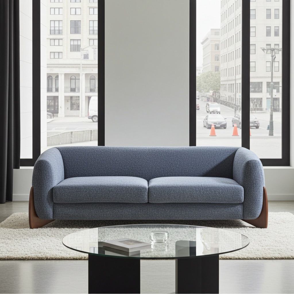 A two-seat sofa upholstered in textured blue fabric with dark wood legs, positioned in a modern living room setting with a glass-top coffee table and large windows showing a city street view.