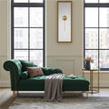A green velvet chaise lounge with gold nailhead trim and gold legs, styled in a bright, modern room with large windows, a gray throw blanket, and a small side table.