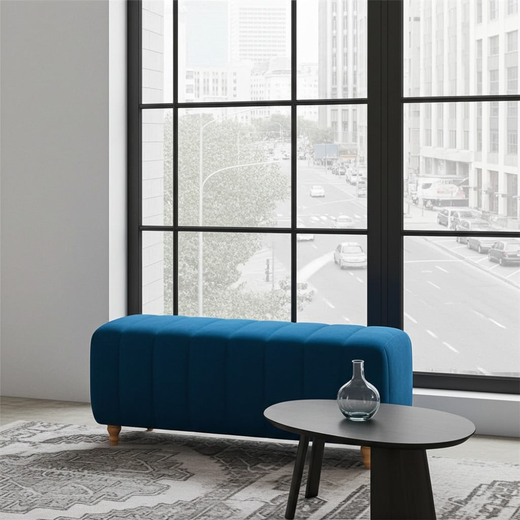 A modern tufted bench upholstered in deep teal blue fabric with light wood legs, positioned in a contemporary room in front of a large black-framed window overlooking a city street.