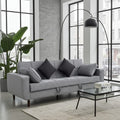 A modern light gray fabric sofa with dark gray throw pillows, styled in a contemporary living room with a glass coffee table, black floor lamp, and a large potted plant.