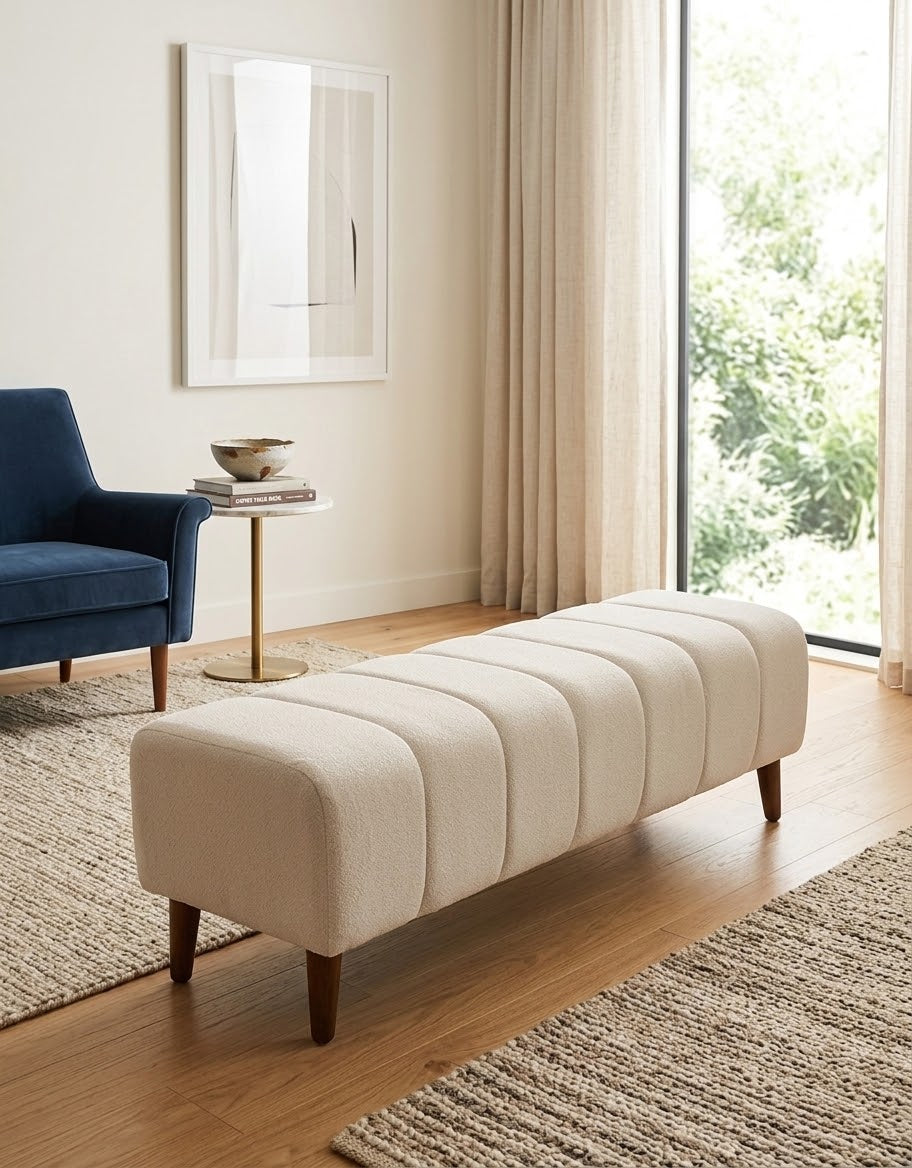 A cream-colored tufted fabric bench with dark wooden legs, styled in a bright, modern living room next to a blue armchair and a large window.