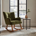 An olive green velvet rocking chair with wooden rockers, positioned next to a small round side table with a vase in a bright room with a large window.
