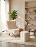 A cream tufted rocking chair with a matching ottoman in a bright, modern living room setting with a wooden bookshelf, rug, and potted plant.