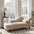 A cream tufted chaise lounge with dark wooden legs sits in a bright, modern living room with large windows, a side table with a lamp, and a patterned rug.